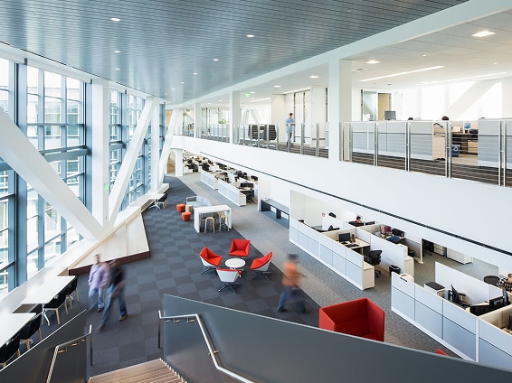 A spacious, modern open-plan office with white desks, red lounge chairs, and large glass windows allowing natural light to flood the space.