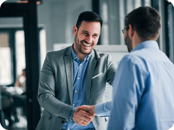 Two business professionals shaking hands in a modern office, with one smiling confidently in a grey blazer.