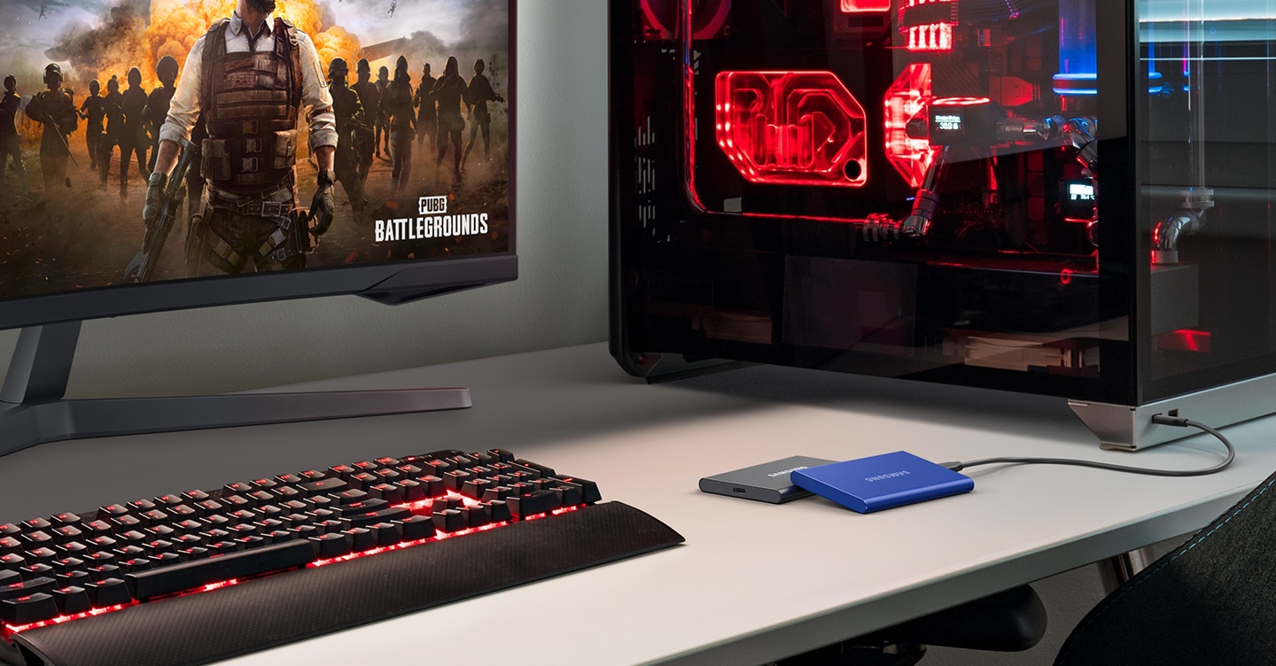 机の上にモニター、キーボード、Samsung T7 ポータブル SSD を備えたゲーム用セットアップ。その横に赤色の内部照明を備えたゲーム用 PC が置かれています。