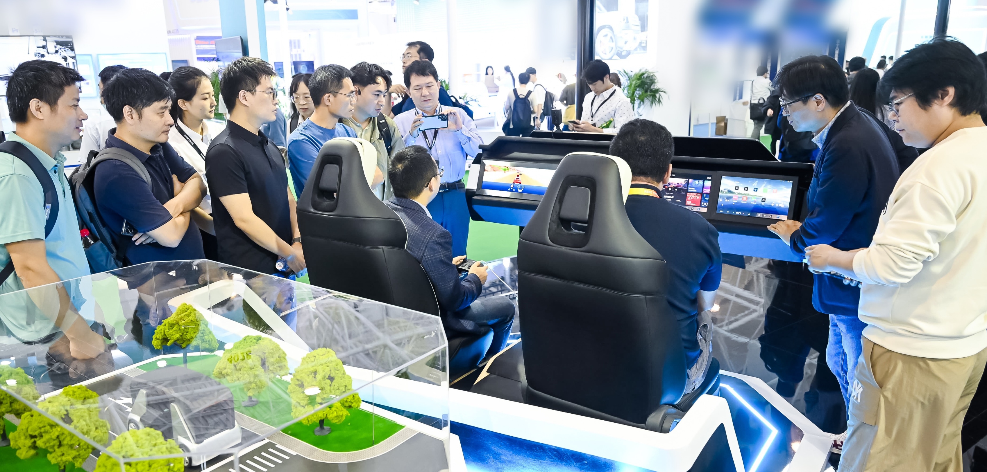 一群人正在展位上与汽车仿真设备互动，包括汽车座椅和仪表盘屏幕，展位上有一个微型道路和汽车模型。