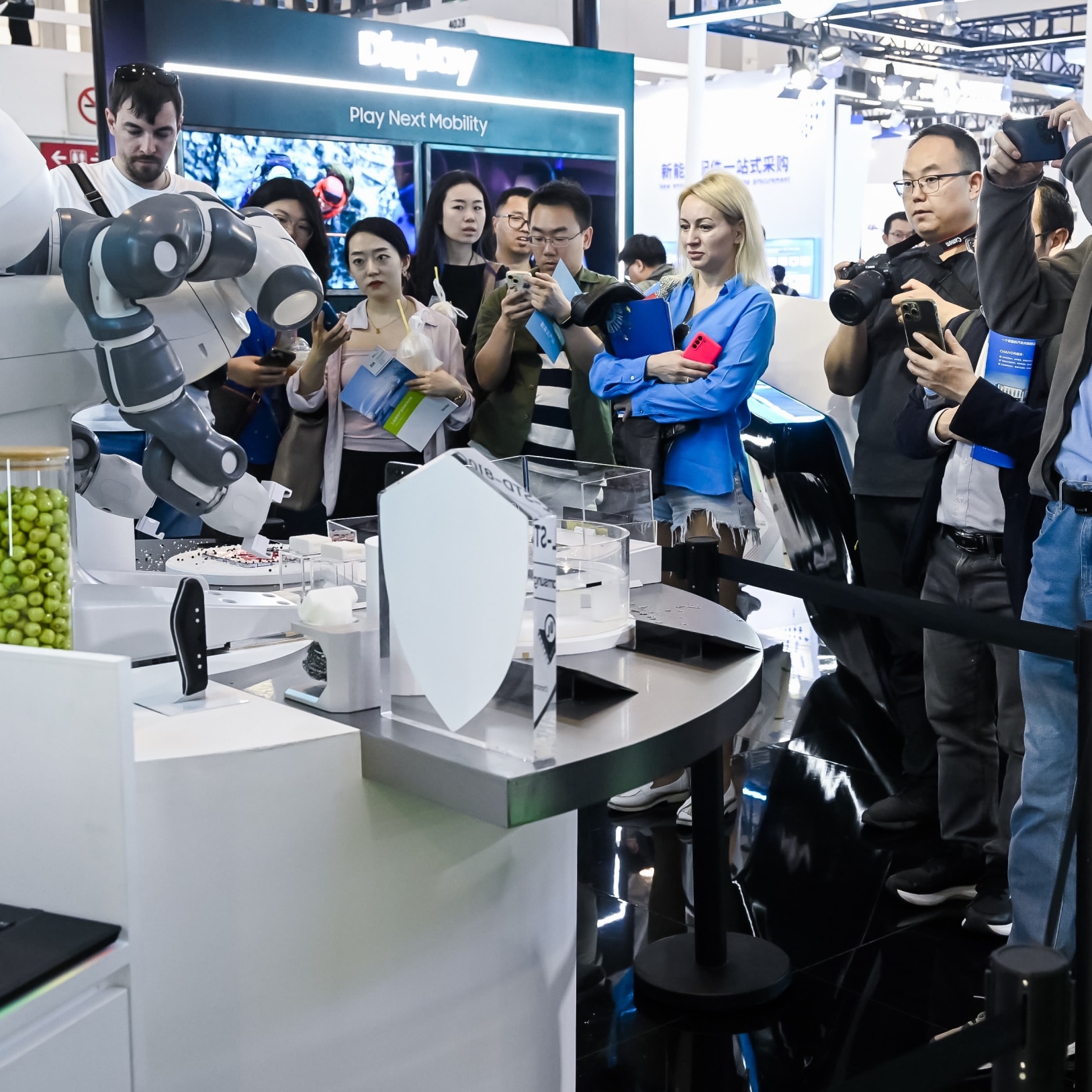 车展的参观者们观看并拍摄一个机器人执行任务，周围有信息展示和数字屏幕。