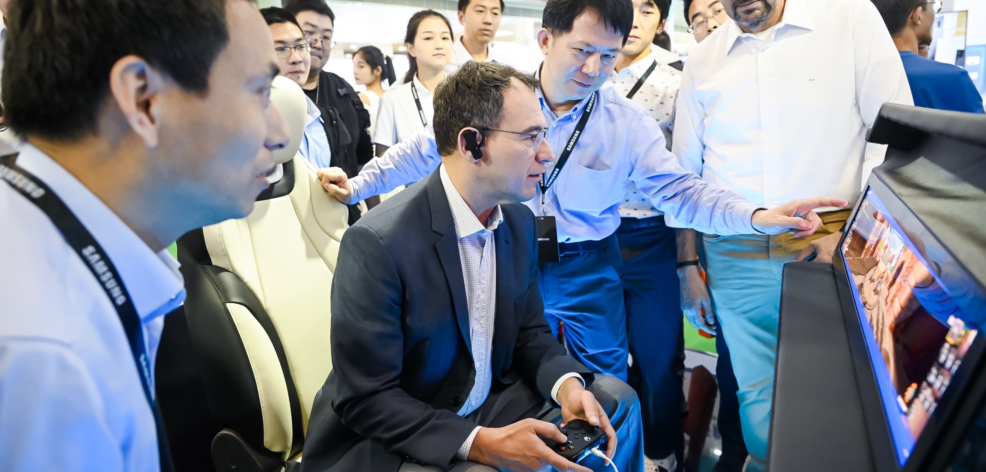 2024年北京车展的一群参观者聚集在一名男子周围，他正在与游戏手柄互动，专注于显示器上的游戏画面。