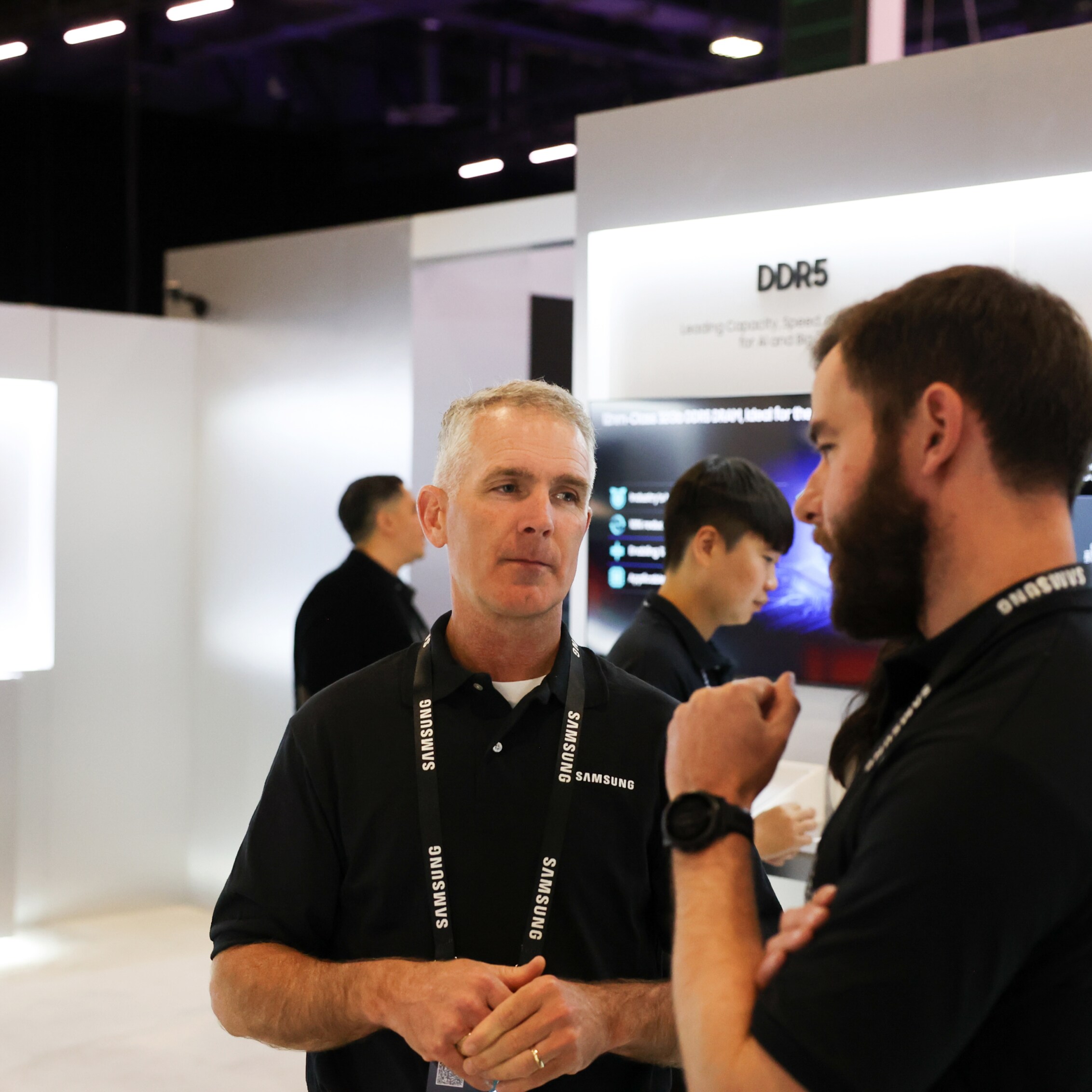 A discussion between two professionals at Dell Technologies World 2024, taking place at the Samsung Semiconductor exhibit surrounded by high-tech displays.