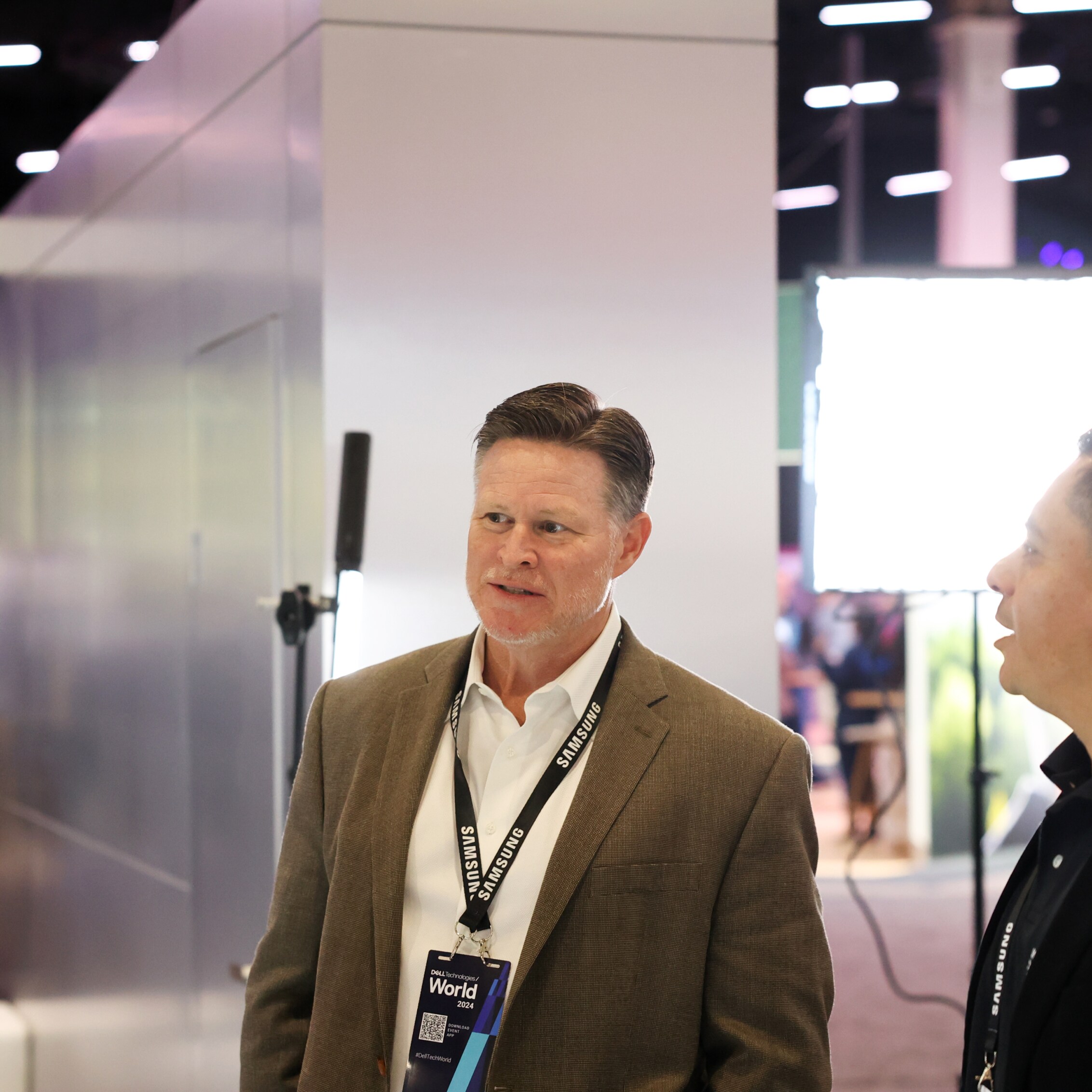 A conversation between two attendees at Dell Technologies World 2024, taking place near a booth with vibrant event lighting in the background.
