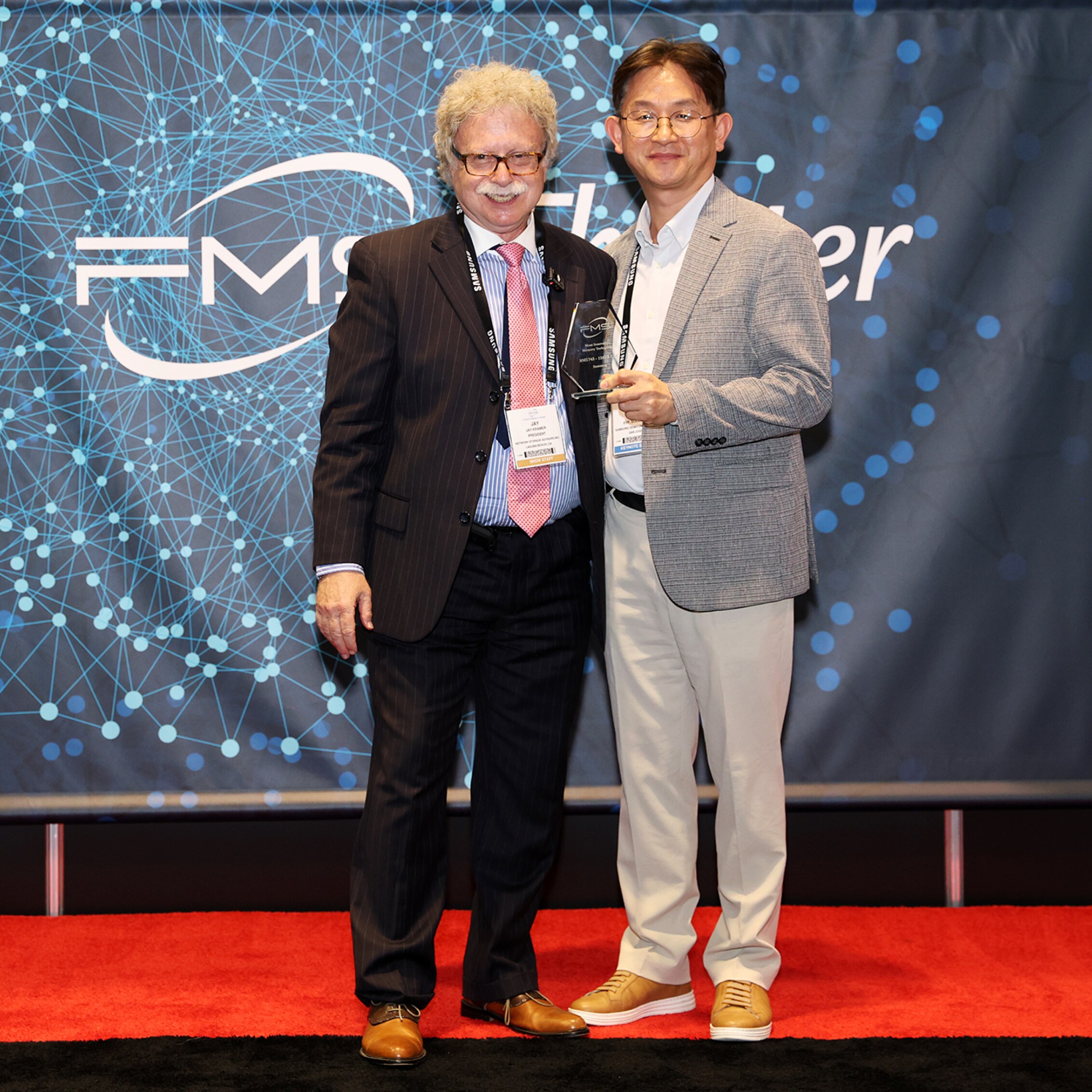 Two men posing with an award on stage at FMS 2024, with a backdrop featuring the FMS logo and a red carpet beneath them.