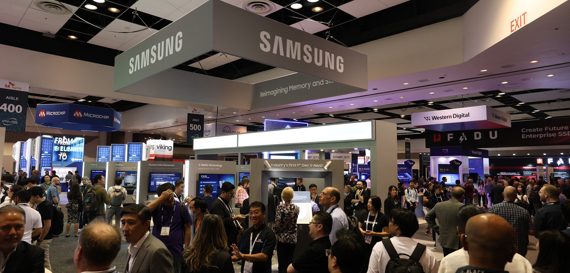 A bustling crowd at FMS 2024 exhibition, with large Samsung banners hanging from the ceiling and various booths visible.