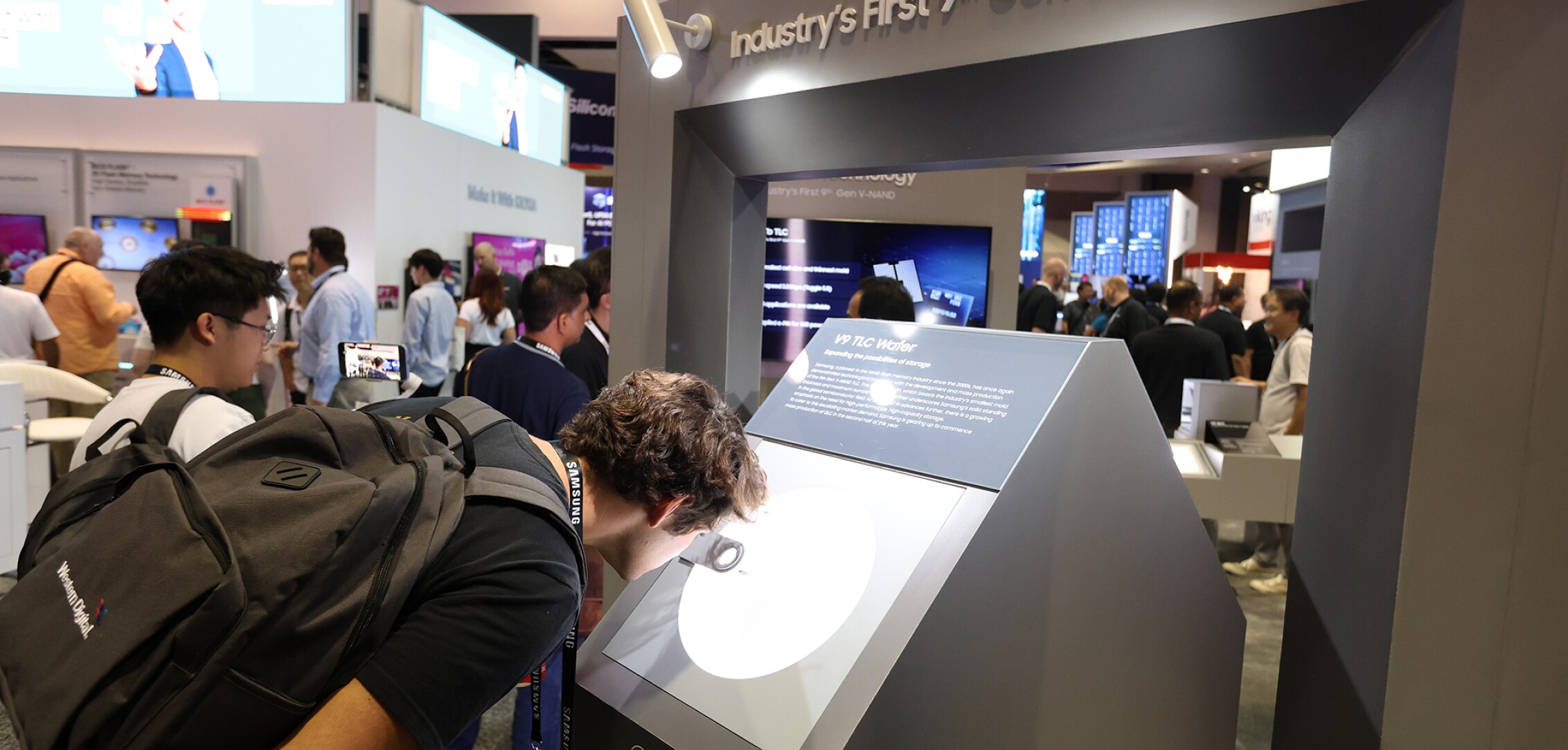 An attendee closely examining the Samsung 9th Gen V-NAND display at the Samsung Semiconductor FMS 2024, with others exploring nearby exhibits.