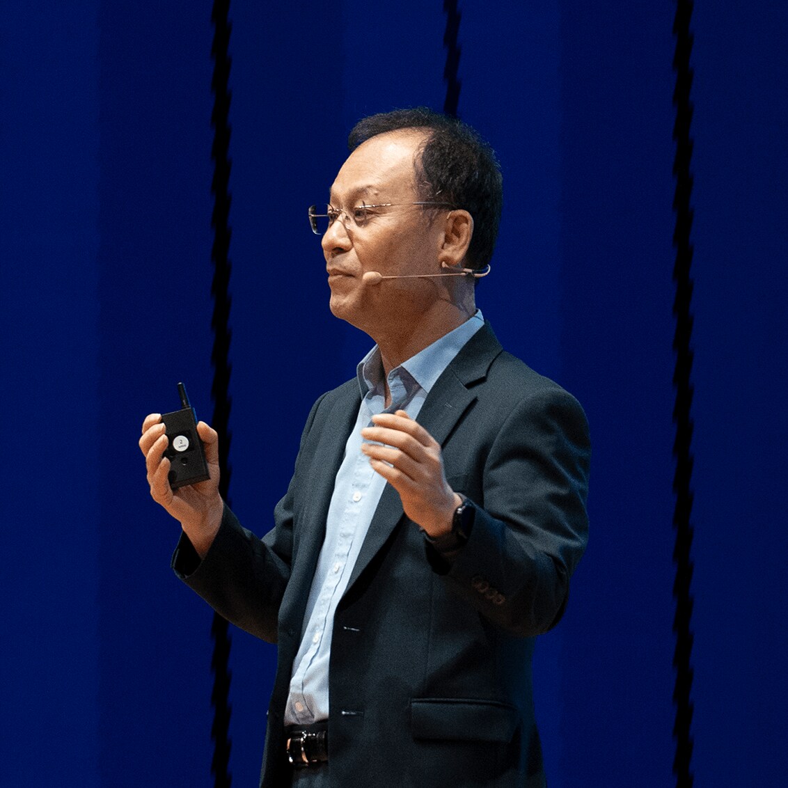 A speaker in a dark suit delivers a presentation on stage against a backdrop of vertical blue stripes, holding a clicker and gesturing with his hands.