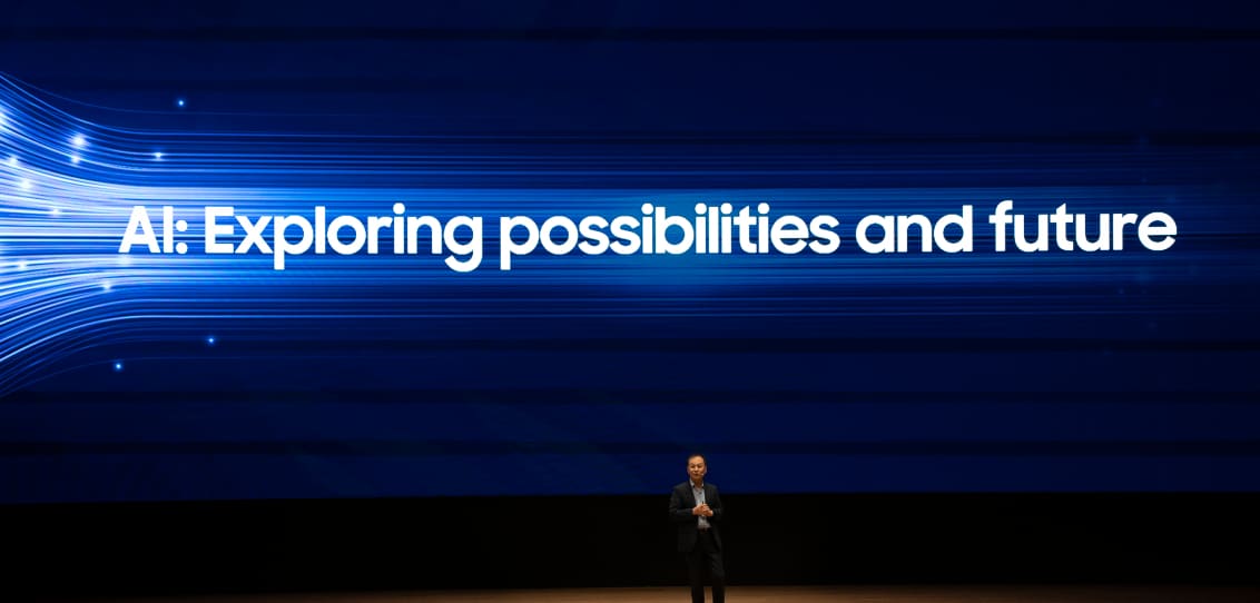 A presenter on stage with a large blue screen displaying the text 'AI: Exploring possibilities and future.'