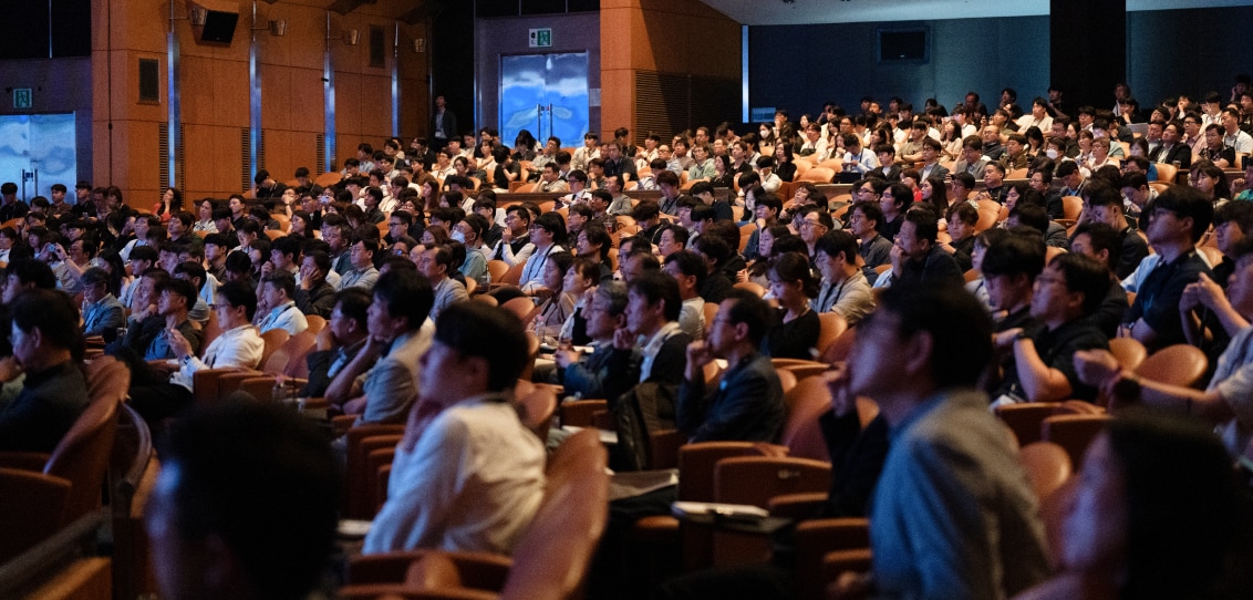 A large audience attentively listening to a presentation in a dimly lit auditorium, with rows of occupied seats extending to the back.