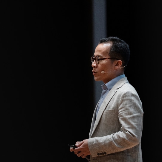 A man in glasses and a light grey blazer giving a presentation on stage, holding a remote with a dark background behind him.