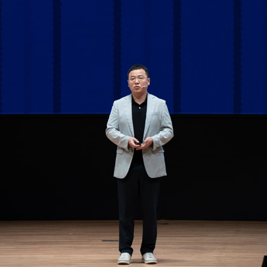 A man in a light grey blazer presents on stage with a large blue screen behind him.