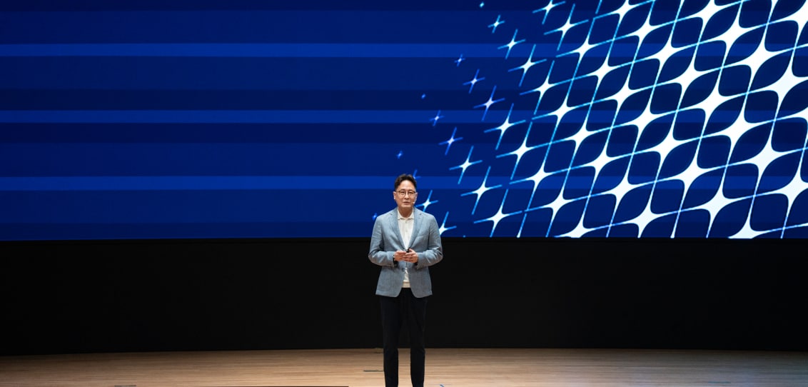 A speaker in casual business attire standing on a stage, with a backdrop featuring blue and white abstract designs.