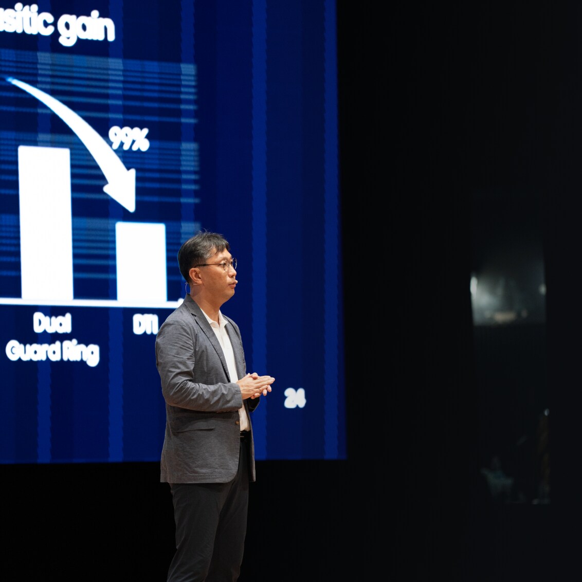 A man in a grey blazer presenting on stage, standing beside a large screen.