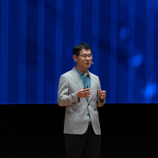 A man in a light grey blazer presenting on stage, with a large blue screen behind him displaying the word 'Opportunities.'