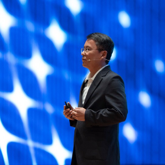 A speaker holding a remote, dressed in a dark suit, presenting in front of a blue background.