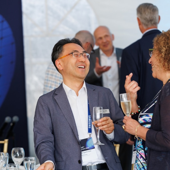 A man in formal attire smiling and holding a champagne glass while engaging in conversation at a networking event