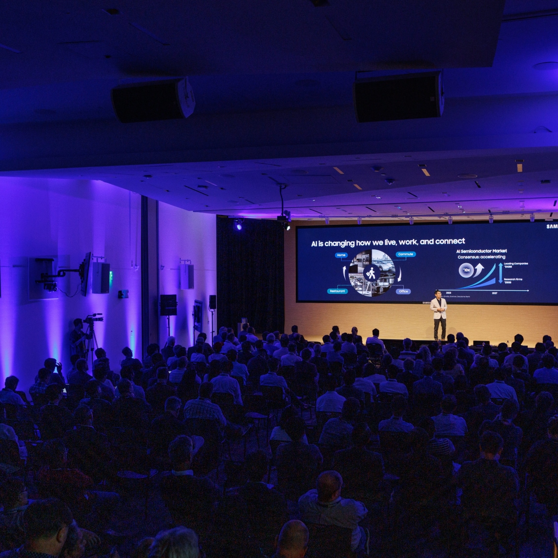 A corporate event showcasing a speaker presenting AI innovations, with blue-themed lighting and a full audience