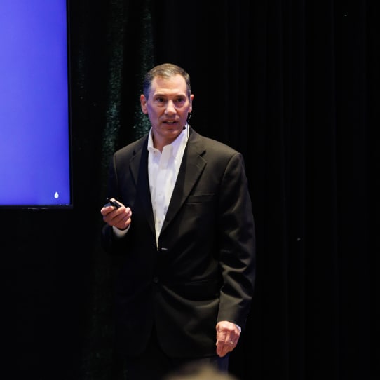 A professional speaker on stage during a conference, with a blue-themed slide partially visible behind him.