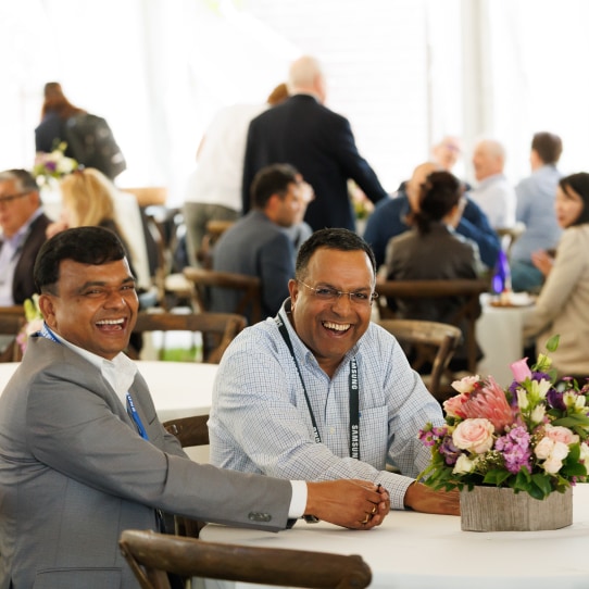 Two men smiling brightly while seated at a table decorated with a flower arrangement during a networking event