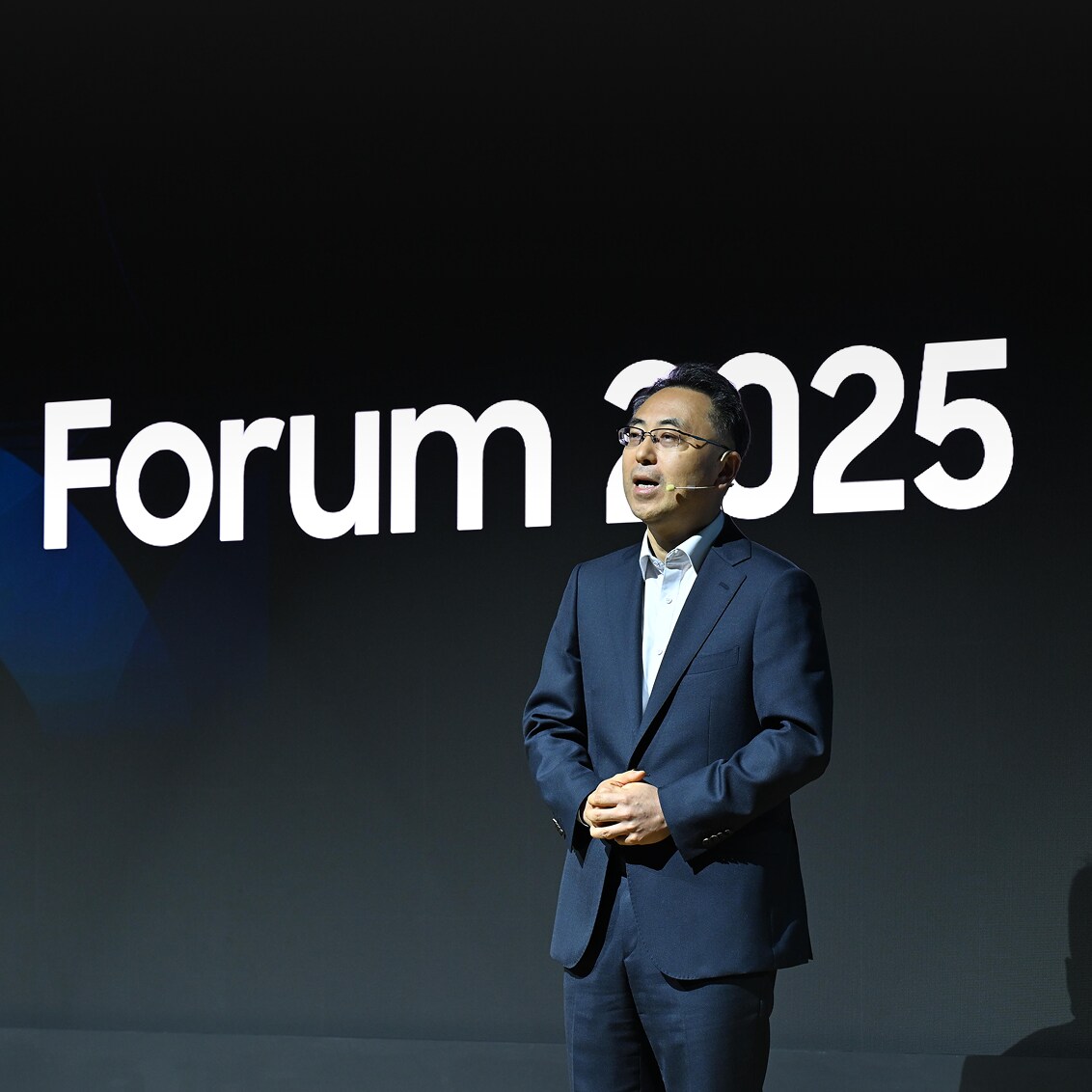 A speaker in a navy suit delivers a keynote address at the 2025 Forum, standing in front of a large digital screen displaying 'Forum 2025.'