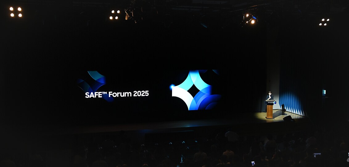 A presenter stands at a podium on the right side of a large stage during SAFE™ Forum 2025, with the event logo displayed prominently on a wide screen.