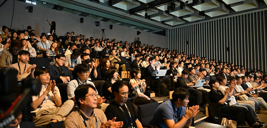 A large audience attentively watches a presentation in a modern auditorium, with many attendees applauding and some using laptops.