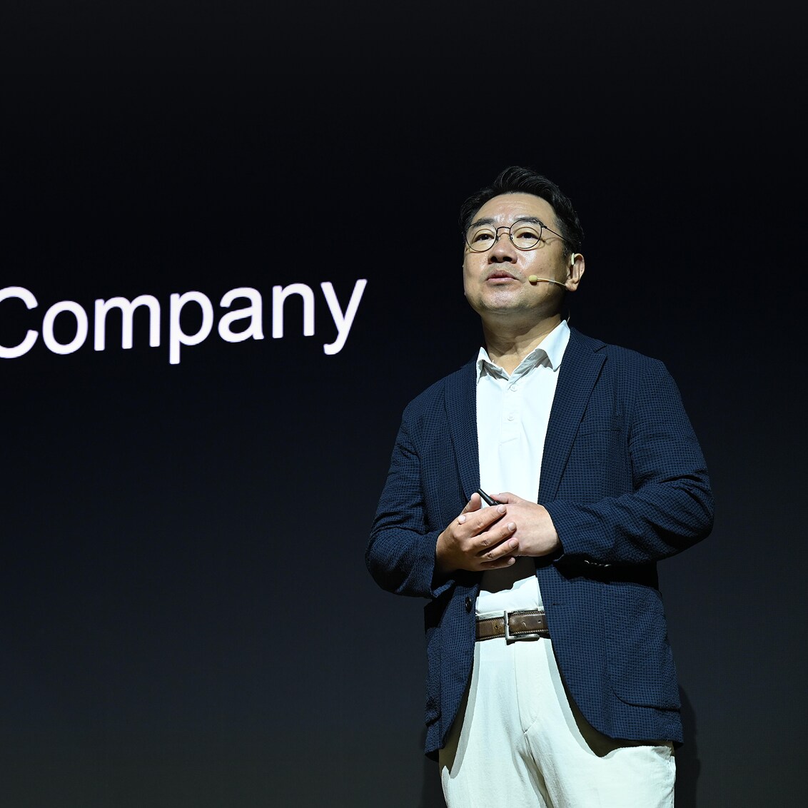 A speaker wearing a headset microphone and navy blazer presents on stage in front of a dark screen partially displaying the word 'Company.'