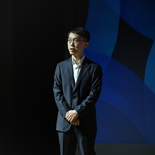 A speaker in a dark suit and white shirt stands under spotlight on stage, with a geometric blue pattern projected behind him.