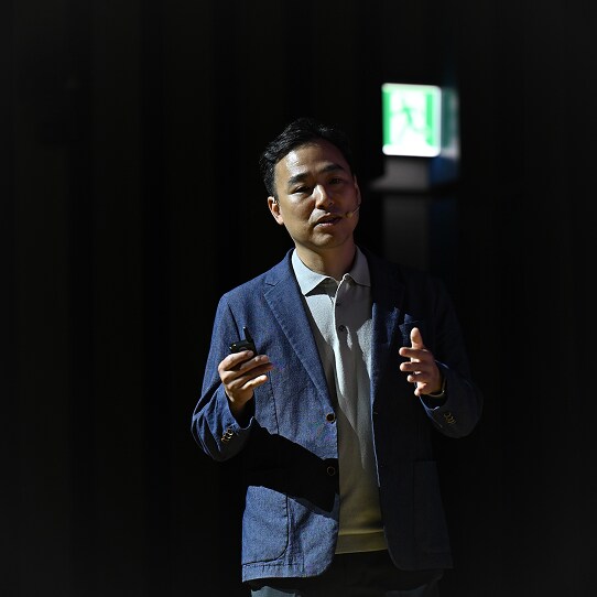 A presenter in a blue blazer and headset microphone gestures during a talk on a dimly lit stage.