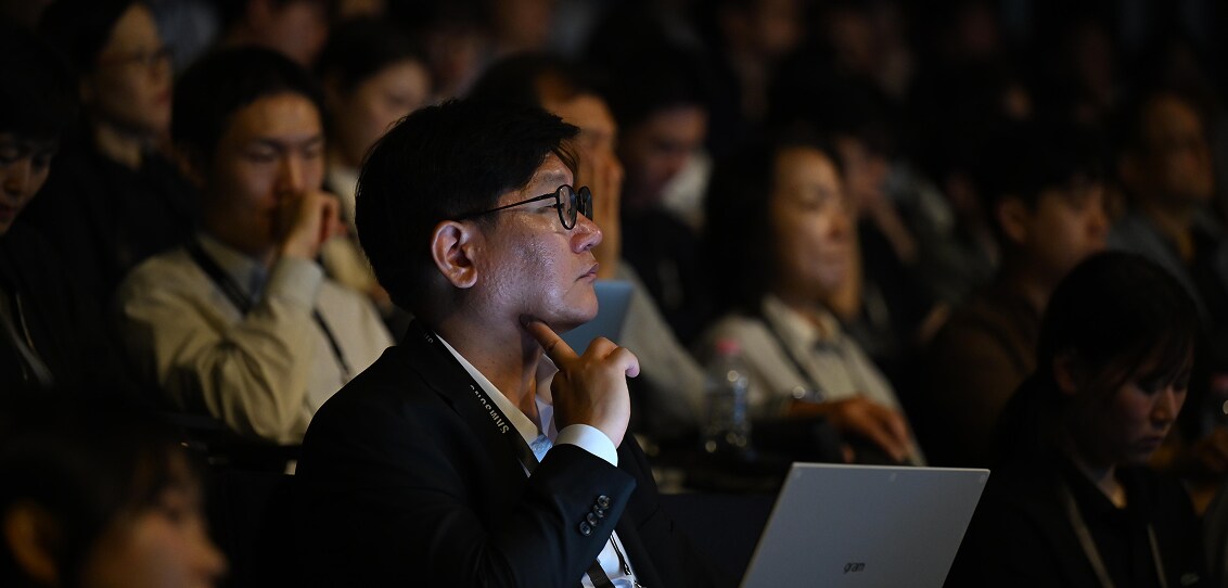 A focused attendee listens intently during a conference, resting his chin on his hand, surrounded by others engaged in the session.