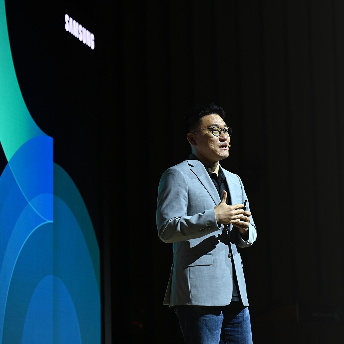 A Samsung presenter in a light gray blazer speaks on stage, standing beside a vibrant screen with blue and green abstract graphics.