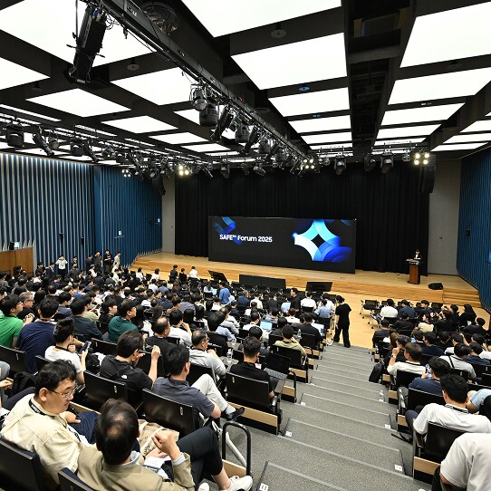 A wide view of the SAFE™ Forum 2025 shows a large audience seated in a modern auditorium, attentively watching a speaker on stage.