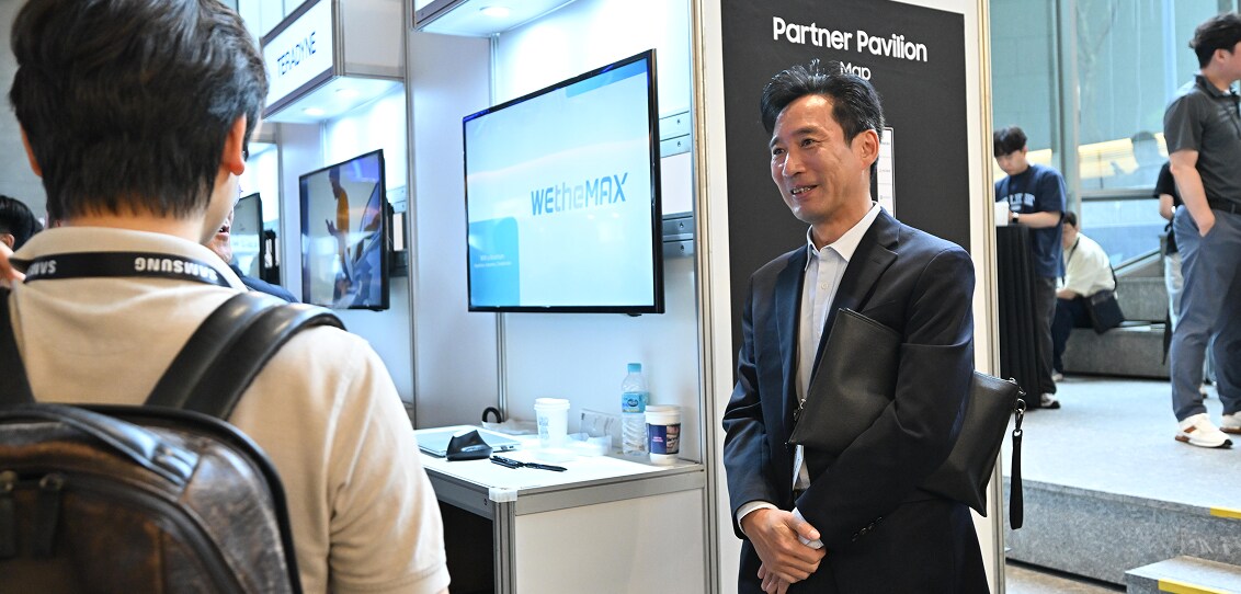 A business representative at the Partner Pavilion booth speaks with an attendee during SAFE™ Forum 2025, with digital displays and promotional materials in the background.