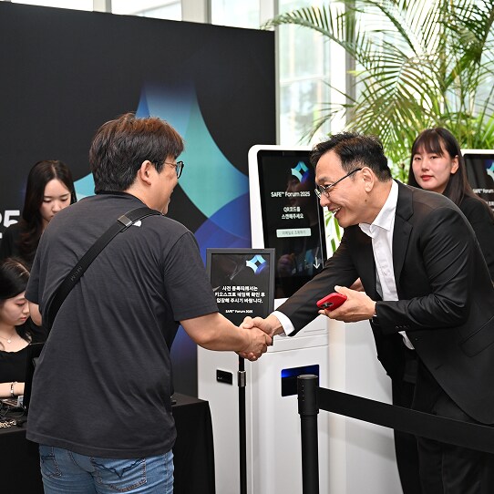 A representative warmly greets an attendee with a handshake at the SAFE™ Forum 2025 entrance, in front of self check-in kiosks.