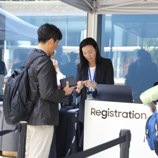 A registration staff member assists a participant at the SAFE™ Forum 2025 booth, handing out a badge under a canopy setup.