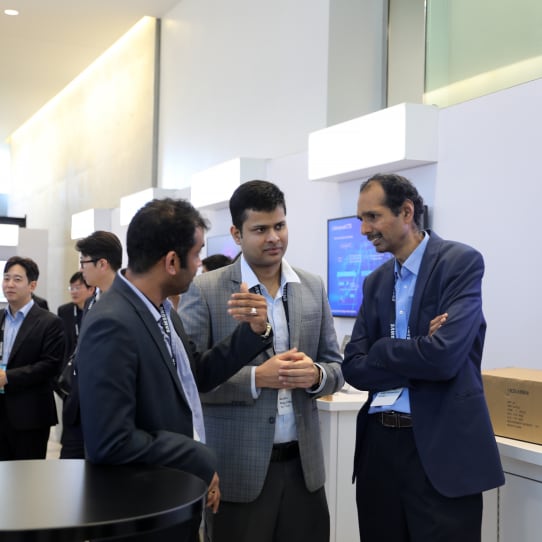 Three men in business attire converse during a networking session at the SAFE™ Forum 2025, with informational screens in the background.