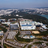 An aerial view of a large semiconductor facility complex, surrounded by roads, greenery, and urban development in the distance.