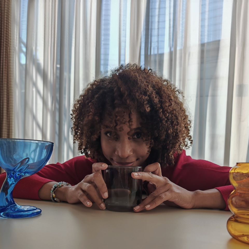 Side-by-side comparison image showing a woman with curly hair sitting at a table, with noticeable differences in brightness and color between the left and right sides.