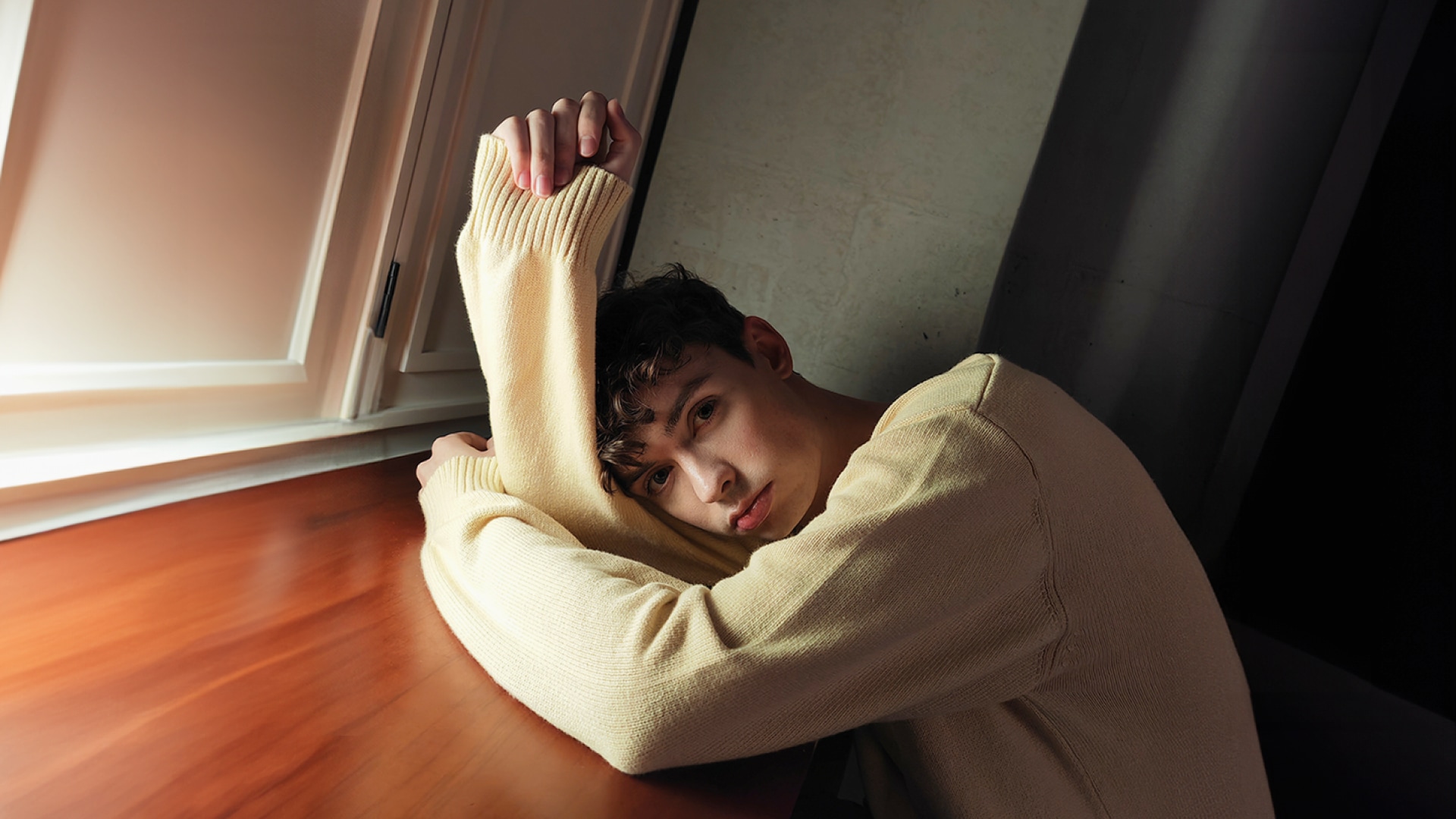 Photo of a person wearing a cream sweater, resting their head and arm on a wooden table, in a dimly lit room that only casts a faint light.