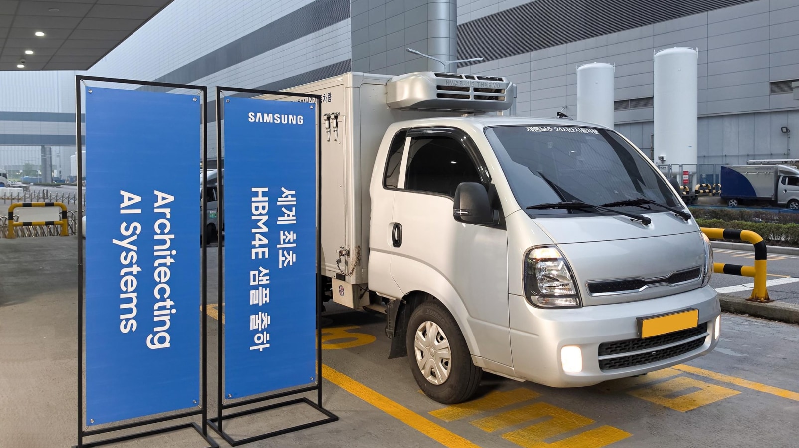A refrigerated truck parked next to blue signs that read "세계 최초 HBM4E 샘플 출하" and "Architecting AI Systems" from Samsung.