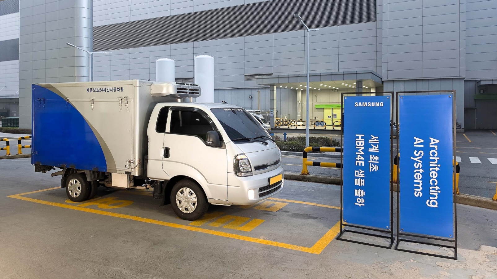 A refrigerated truck parked next to blue signs that read "세계 최초 HBM4E 샘플 출하" and "Architecting AI Systems" from Samsung.