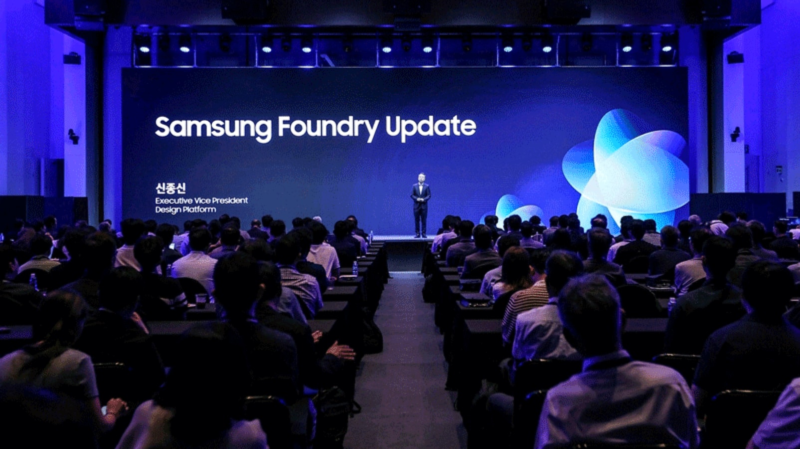 검은색 정장을 입은 발표자가 "Samsung Foundry Update"라는 문구가 표시된 대형 스크린 앞 단상에 서 있고, 많은 참석자들이 긴 테이블에 앉아 무대를 바라보고 있습니다.