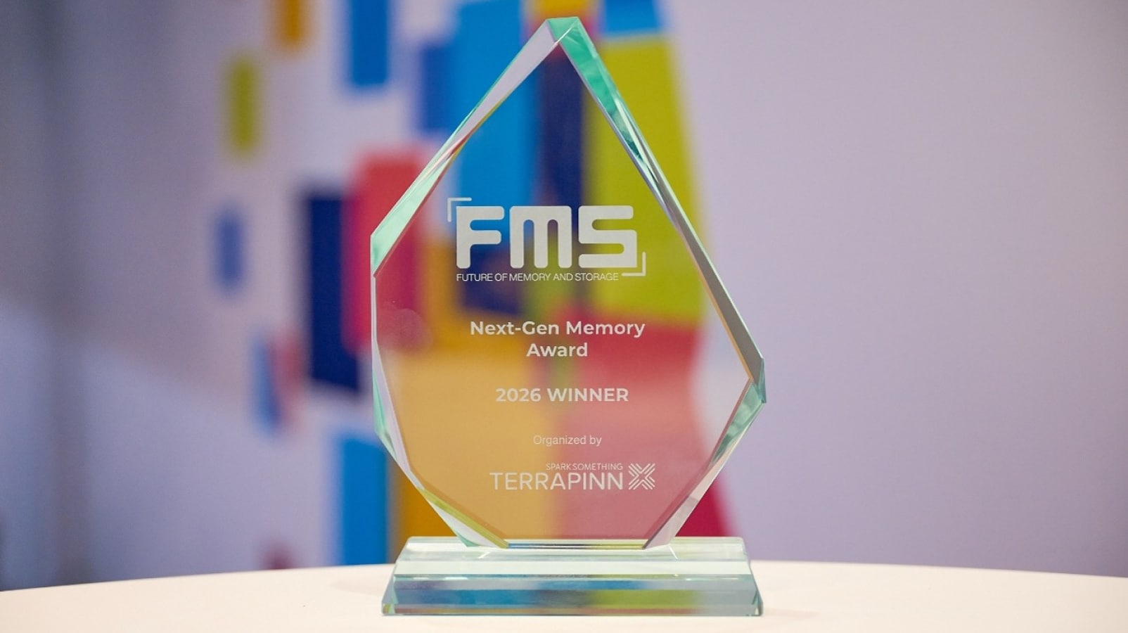 흰색 표면 위에 놓인 유리 소재의 수상 트로피로, 'FMS Next-Gen Memory Award 2026 Winner'라는 문구가 새겨져 있으며 배경에는 색색의 패널이 흐릿하게 보입니다.