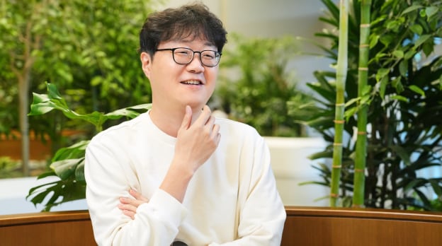 센서 PA 팀의 TL이 실내 녹지 공간에서 턱에 손을 올린 채 미소를 지으며 인터뷰 중인 모습.