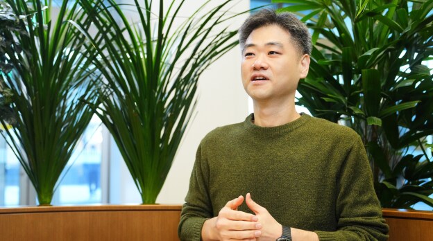 Security&Power제품개발팀의 강현재 TL이 녹색 니트 스웨터를 입고 식물로 채워진 사무 공간에서 여유로운 표정으로 이야기하는 모습