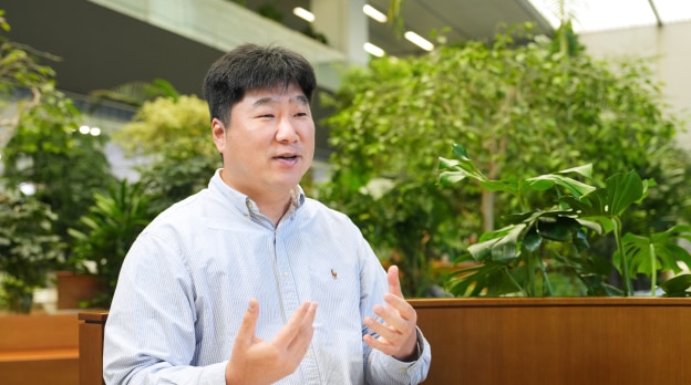 Security&Power제품개발팀의 이인 님이 밝은 표정으로 손짓하며 설명하고 있는 모습으로, 녹색 식물이 있는 실내 공간에서 대화하는 분위기