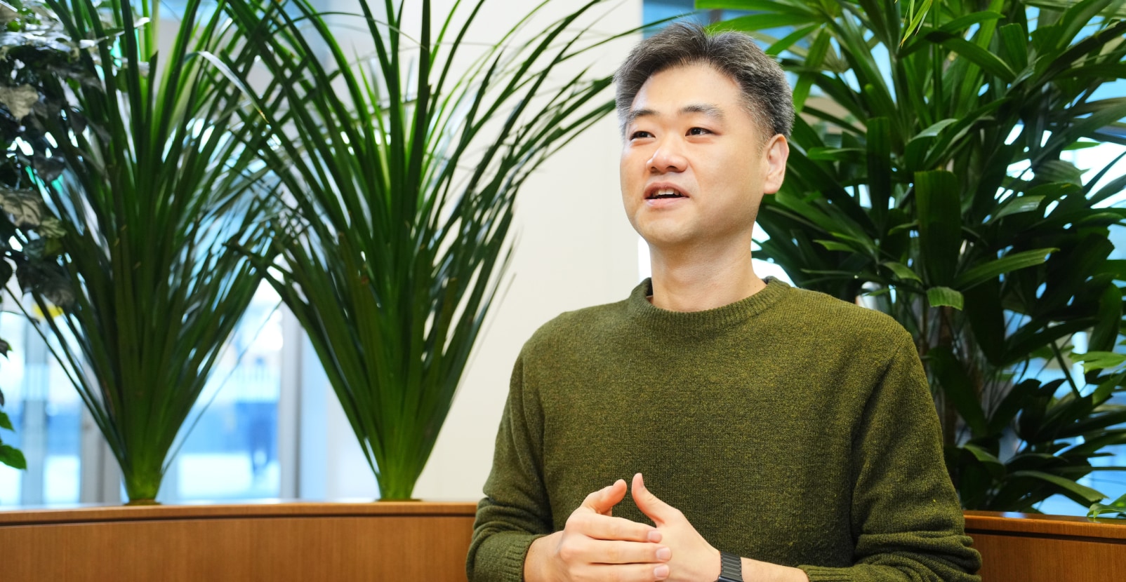 Security&Power제품개발팀의 강현재 TL이 녹색 니트 스웨터를 입고 식물로 채워진 사무 공간에서 여유로운 표정으로 이야기하는 모습