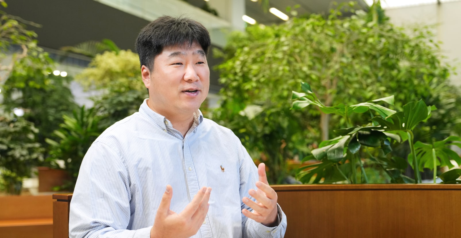 Security&Power제품개발팀의 이인 님이 밝은 표정으로 손짓하며 설명하고 있는 모습으로, 녹색 식물이 있는 실내 공간에서 대화하는 분위기