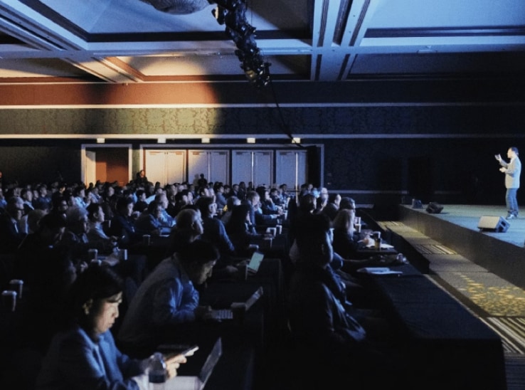 A presenter speaks to an audience in a dark conference hall, with attendees seated and facing a brightly lit stage.